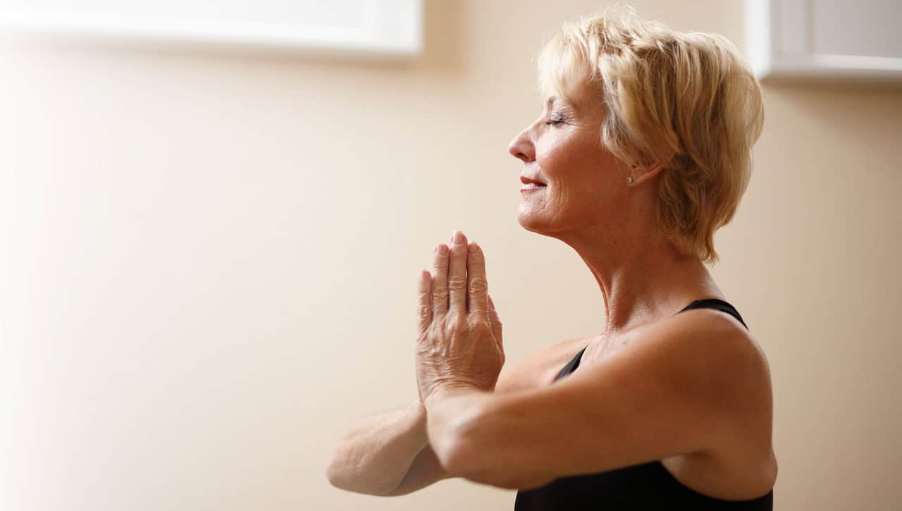 A smiling older woman with her eyes closed and hands pressed together, breathing calmly