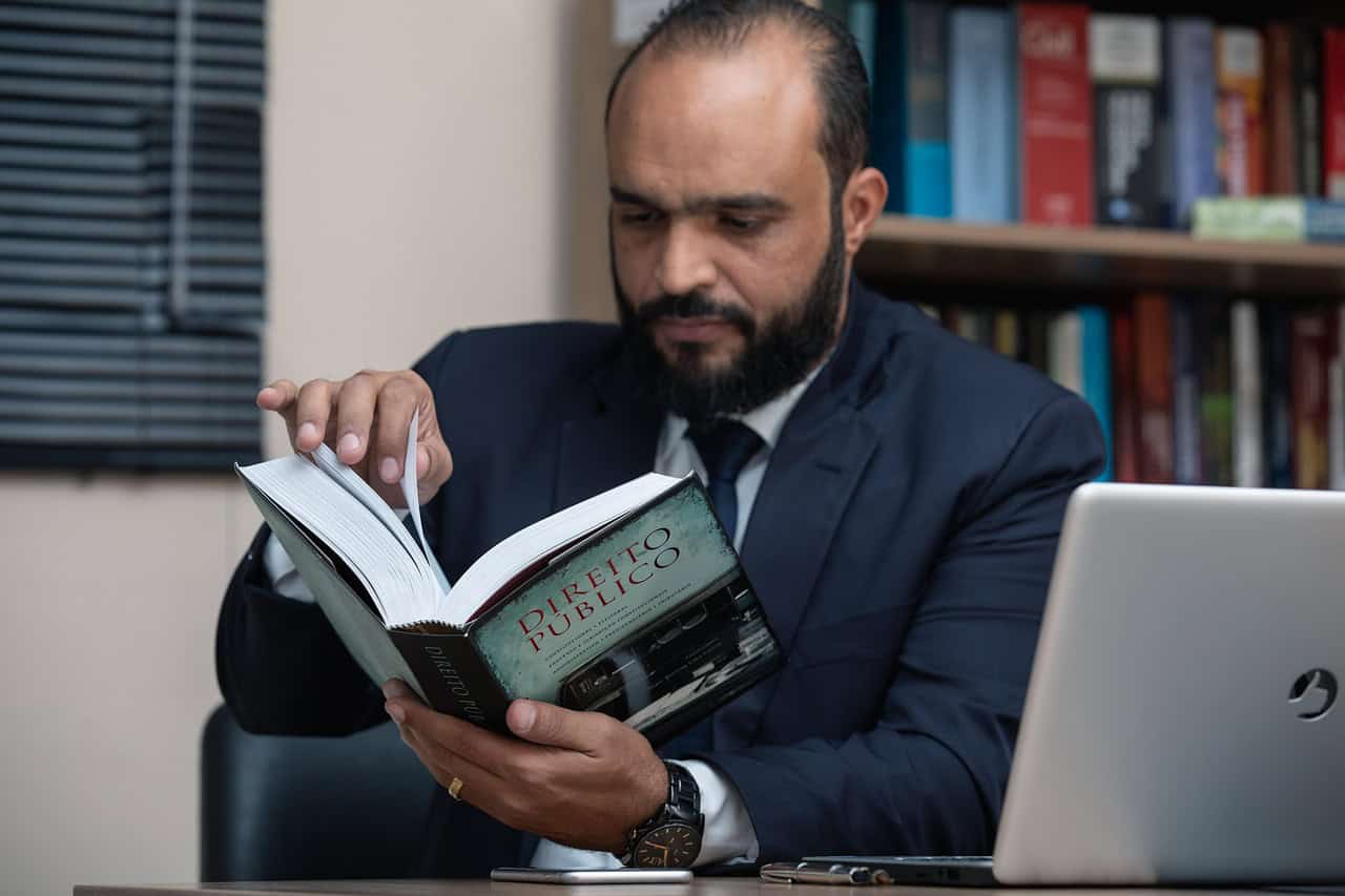 A man reading a book at his desk next to a laptop