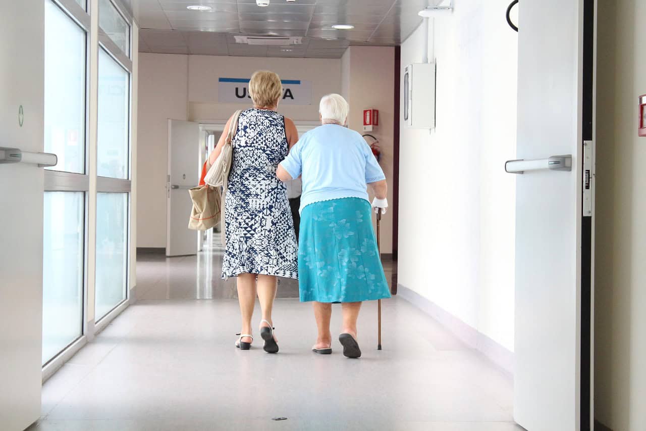 A caregiver walking arm in arm with an older woman down a bright hallway