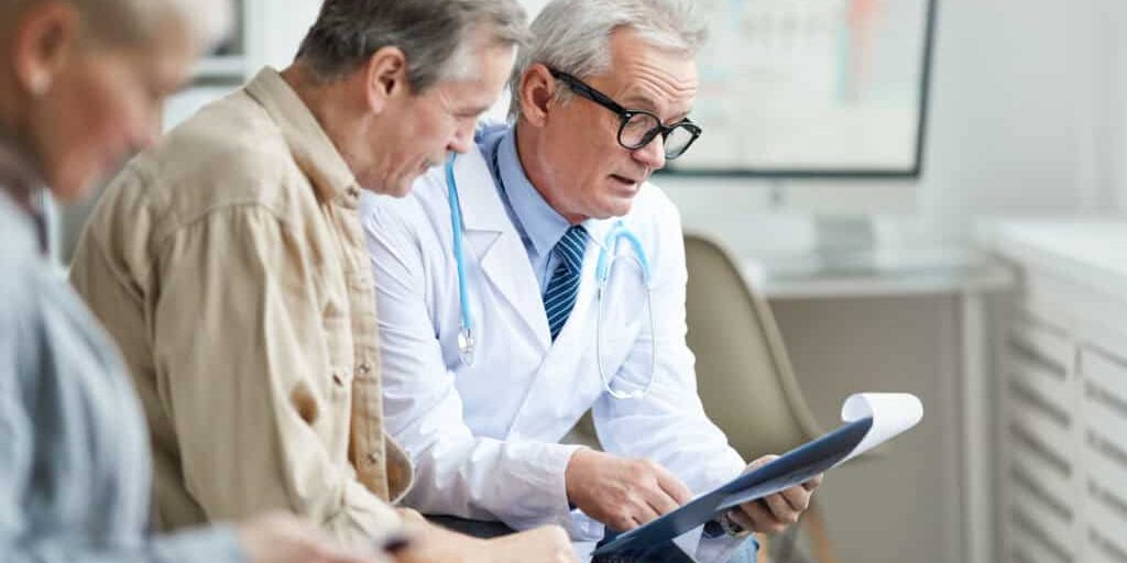 Senior male doctor in glasses holding clipboard with papers and commenting test results while talking to mature patient